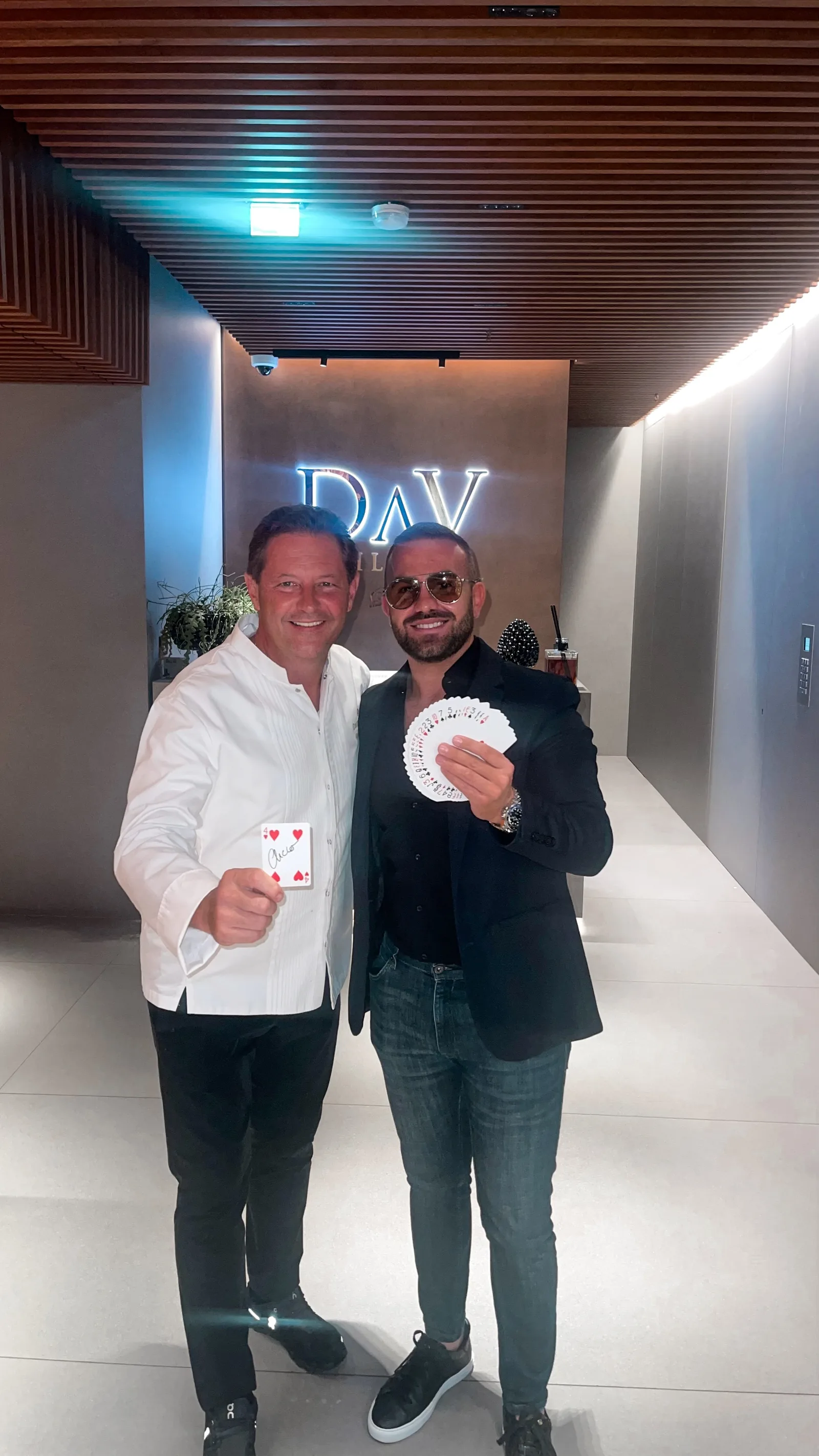 Chef Enrico Cerea of the three-Michelin-starred Da Vittorio holding a signed four of hearts beside Matteo Cammisa, who fans a deck of cards, under the illuminated DaV sign in Milan