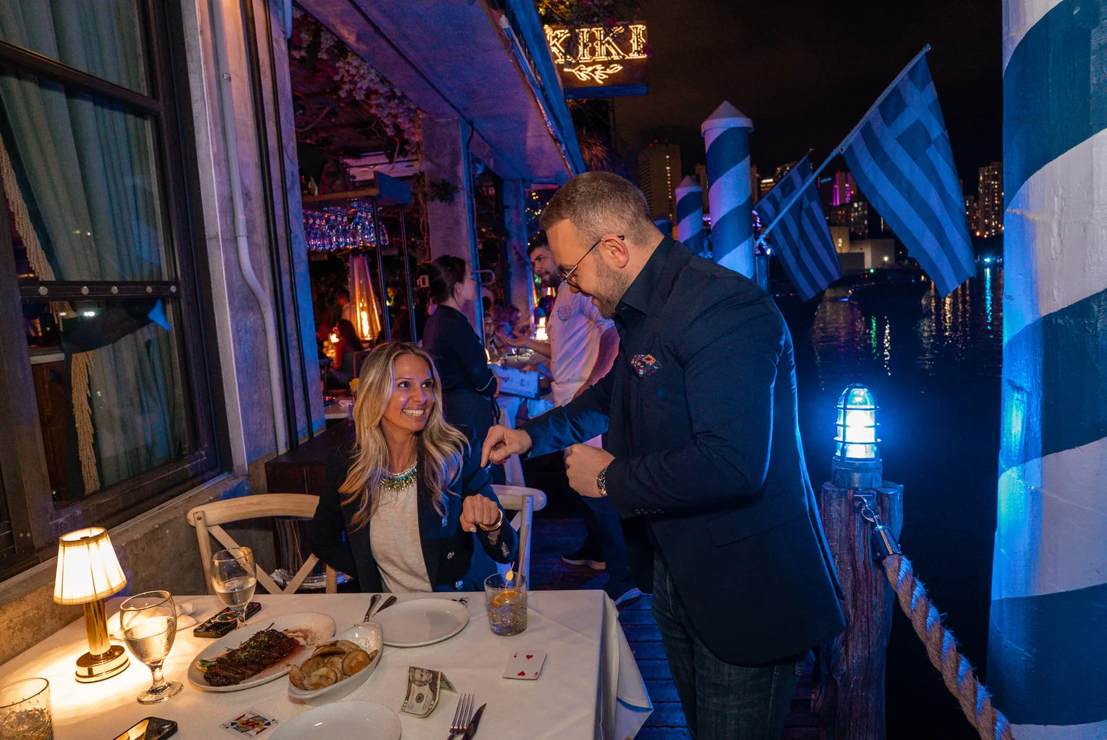 Matteo Cammisa performing tableside at Kiki on the River, with the KIKI sign and Greek flags over the Miami River behind him